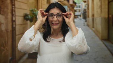 Beyaz bluzlu kadın gözlüğü iki eliyle düzeltirken güneşli, dar bir kasaba caddesinde gülümsüyor..