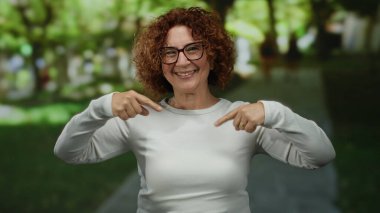 Güneşli bir parkta gülümseyen ve el kol hareketi yapan bir kadın, açık havada, gözlük takıyor ve günlük beyaz kıyafetler giyiyor, bulanık yeşillik ve ağaçlarla çevrili, neşeli bir anı yakalıyor..