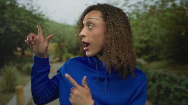 Ağzı ve eli açık bir kadın ormandaki yeşil yaprakların arasında kirli bir patika boyunca yan tarafı işaret ediyor; merak..