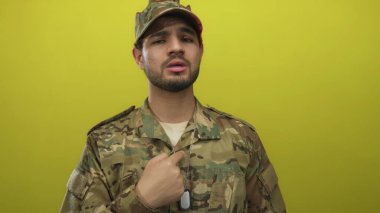 Kamuflaj üniformalı genç İspanyol asker ciddi bir ifadeyle sarı stüdyoda parmak göğüse işaret ediyor..