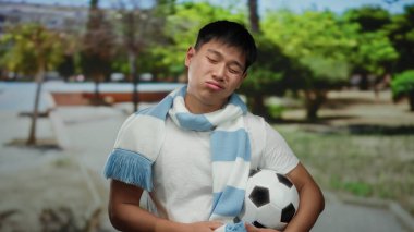 Parkta elinde futbol topuyla gezen ve eşarp takan genç bir adam, güneşli bir açık havada uykulu görünüyor, arka planda yeşil ağaçlar var..