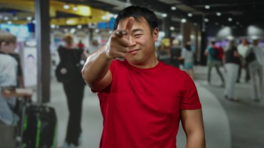 Kırmızı gömlekli genç adam havaalanı terminalinde el hareketi yapıyor. Bulanık gezginler ve parlak ışıklandırmayla seyahat mizahı anlayışı aktarıyor..