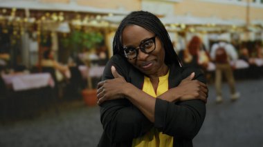 Açık hava pazarında kollarını kavuşturmuş gülümseyen bir kadın, şehir atmosferini sıcak bir ifadeyle harmanlıyor, gözlük takıyor ve sıradan bir kıyafet giyiyor.