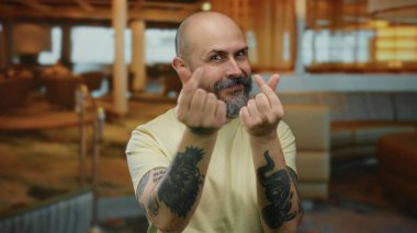 Sakallı, kel bir adam şık bir otelde parmaklarını kalp yapıyor. Kapalı bir odada, arka planda bulanık oturma alanı olan dövmeler yapıyor. Rahat bir ortam yakalıyor.