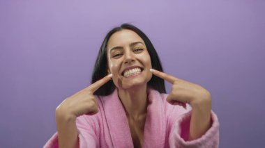 Pembe bornozlu genç İspanyol kadın gülümsüyor ve mor arka planda dişlerine işaret ediyor. Mutluluk ve diş bakımı taşıyor..