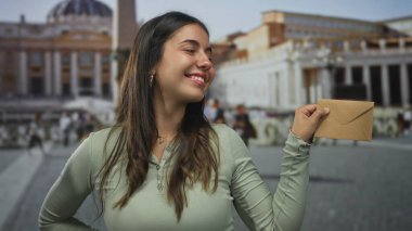 Vatikan 'da St. Peter Meydanı' nda elinde bir zarf tutarken gülümseyen, İtalya 'da canlı ve neşeli bir doğa deneyimi sergileyen bir kadın..