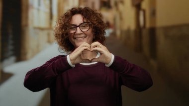 Kıvırcık saçlı ve gözlüklü bir kadın şehir arka planında gülümserken kalp hareketi yapıyor, dışarıda mutluluk ve pozitifliği ifade ediyor..