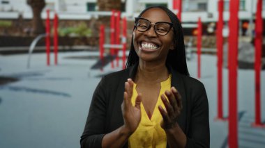 Dışarıdaki spor salonunda kırmızı ekipmanlarla gülümseyen bir kadın. Gözlüklü, sarı tişörtlü, gündelik tarzda ve güneşli gökyüzünün altında neşeli bir ifade. Mutluluk ve sıcaklığı yansıtıyor..