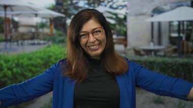 Yüzü, gözlüğü ve kolları açık, mavi ceketli, gülümseyen orta yaşlı bir kadın..