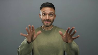 Elleri gri arka planda, sakin hareketler yapan İspanyol bir adam, sıradan yeşil bir kazak giyiyor, tarafsız bir ortamda sükunetini ve güvenini ifade ediyor..