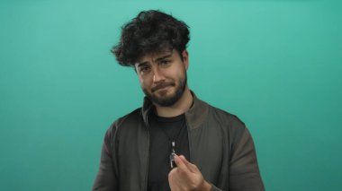 Sakallı ve üzgün yüzlü genç İspanyol bir adam canlı yeşil bir arka plana karşı poz veriyor, günlük kıyafetler ve kolye giyiyor, duyguları ve tepkileri vurguluyor..