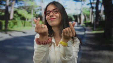 Genç gülümseyen İspanyol kadın, güneşli ağaçlı yolda parmaklarıyla kalp hareketi yapıyor..