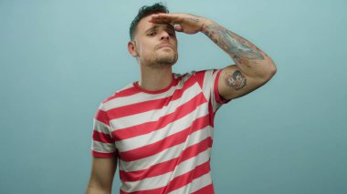 İspanyol erkek dikkatle uzaklığa bakıyor, saf mavi bir arka plana karşı duruyor, dövmelerini ve kendinden emin duruşu olan günlük çizgili gömleğini vurguluyor..