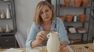 Orta yaşlı kadın sol eliyle kil vazoyu tutuyor ve stüdyoda sağ eliyle çember boyuyor..