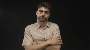 Gözlüklü İspanyol genç adam stüdyoda siyah ışık altında kollarını çaprazlıyor..