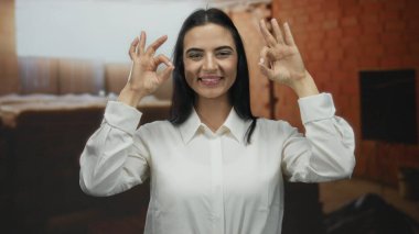 Beyaz tişörtlü genç İspanyol kadın, kırmızı tuğla duvarlarla çevrili kapalı bir inşaat alanında kendine güveni ve pozitifliği olan bir jest yapıyor..