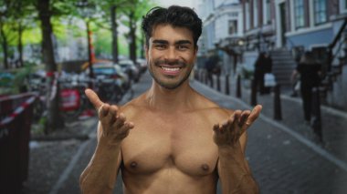 Gömleksiz genç İspanyol adam gülümsüyor ve çıplak göğsü olan şehir sokağındaki kamerayı işaret ediyor..