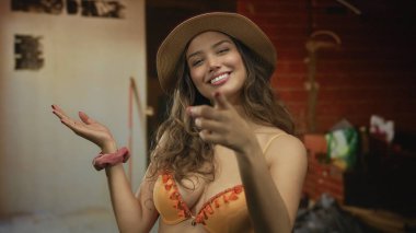 Turuncu bikinili genç İspanyol kadın, hasır şapkalı ve gülümseyen bir inşaat binasında başparmak kaldırıyor..