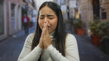 Umutlu bir ifade ve elleri sımsıkı bağlı, şehir binaları ve bitkilerle çevrili, sokakta dua eden bir kadın..