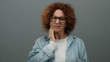 Yüzü gri duvara dönük, acı çeken, orta yaşlı, gözlüklü, kıvırcık saçlı, günlük gömlekli, izole edilmiş arka planda rahatsızlık gösteren bir kadın..