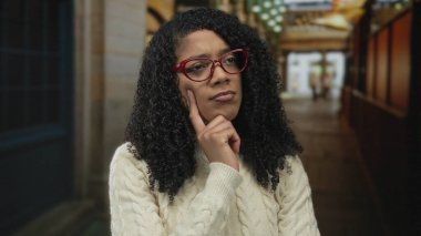 Yumuşak ışık altında düşünmek için çeneye dokunan kırmızı gözlüklü kadın; merak düşüncesi.