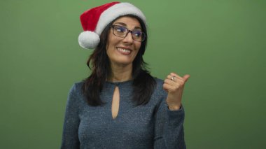Orta yaşlı, gülümseyen, Noel Baba şapkalı ve gözlüklü bir kadın yeşil stüdyoyu işaret ediyor..