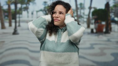 Süveter giymiş İspanyol bir kadın sokakta elleri başının üstünde duruyor, hayal kırıklığına uğramış ya da endişeli görünüyor, ağaçların ve kaldırımların arka planına karşı kurulmuş..