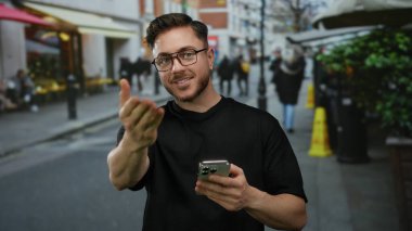 Sakallı genç adam canlı şehir caddesinde telefonu tutarken kameraya el hareketi yapıyor. Kendine güveni tam ve açık havada hareketli bir ortam yaratıyor..