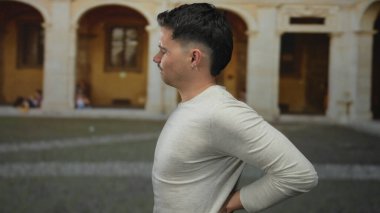 İspanyol bir adam eski bir üniversite ortamında sırt üstü yatıyor, arka planda kemerler olan günlük kıyafetler giyiyor, rahatsızlık ve gerginlik taşıyor..