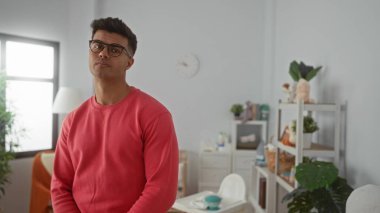 Gözlüklü ve kırmızı kazaklı genç adam, modern iç mekan yaşam tarzını ön plana çıkaran bitkiler ve bebek dekoruyla süslenmiş rahat bir yatak odasında düşünceli bir şekilde duruyor..