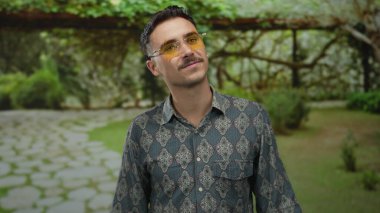 Şık bir gömlek ve yıldız şeklinde güneş gözlüğü takan genç İspanyol bir adam, yemyeşil bir parkta, sarmaşıklarla kaplı arka planda, dışarıdaki manzaranın tadını çıkarıyor..
