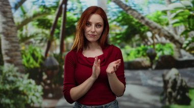 Canlı yeşilliklerle çevrili, kırmızı bir süveter giyen, yemyeşil bir parkta alkışlayan bir kadın, sakin bir açık hava ortamında neşe ve memnuniyeti ifade ediyor..