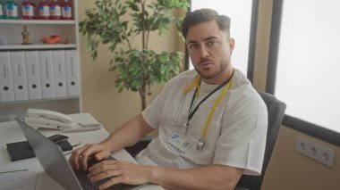 Sakallı genç adam klinik masasında dizüstü bilgisayarda çalışıyor. Steteskopla boynundaki modern iş yeri dekorasyonunu gösteriyor..