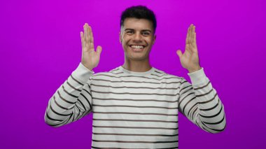 Çizgili tişörtlü, pembe arka planda elleri olan genç İspanyol adam neşeli ve neşeli bir ifade sergiliyor..