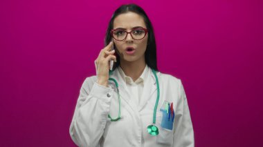 Steteskoplu ve akıllı telefonlu İspanyol kadın doktor canlı pembe bir duvara yaslanmış, tıbbî profesyonelliği ve iletişimi aktarıyor..