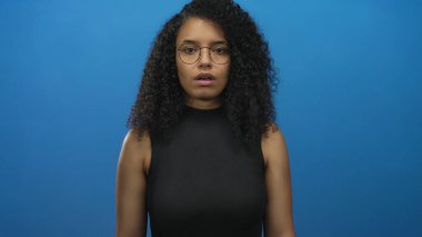 Mavi arka planı gösteren kıvırcık saçlı, gözlüklü, siyah kolsuz bluzlu stüdyo ortamında izole edilmiş, odak ve yön veren bir kadın..