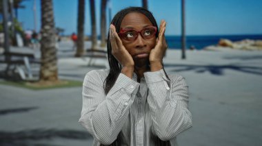 Kıvırcık saçlı, gözlüklü bir kadın güneşli bir sahil kenarında, palmiye ağaçları ve banklarla dolu, rahat bir kumsalda neşeli ifadeler sergiliyor..