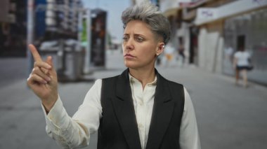 Siyah yelek giyen kısa saçlı kadın, hareketli şehir kaldırımındaki görünmeyen nesneye işaret ediyor; özgüven kararlılığı.