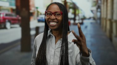 Şehir sokağında gülümseyen kırmızı gözlüklü, kıvırcık saçlı, sahne arkasında neşeli Afro-Amerikan yetişkin sahnesi olan bir kadın..