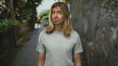 Uzun sarı saçlı ve sakallı bir adam gri tişört giyen taştan duvarın yanındaki şehir sokağında kaşlarını kaldırıyor; belirsizlik.