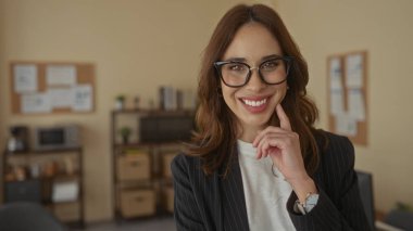 İspanyol kadın kendine güvenerek ofis ortamında gülümsüyor, gözlük ve spor ceket takıyor, modern bir iş ortamında profesyonelliği ve pozitifliği aktarıyor..