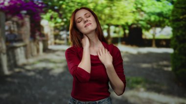 Güneşli bir şehir caddesinde dışarıda duran bir kadın kızıl saçlı ve günlük kıyafetleriyle boyun ağrısı çekiyor. Arka planda bulanık yeşillik ve canlı çiçeklerle çevrili.