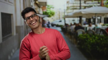 Kırmızı kazaklı yakışıklı bir adam şehir caddesindeki terasta gülümsüyor, sıcaklık ve cazibe saçıyor..