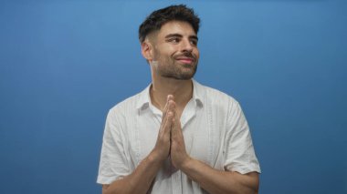 Beyaz keten gömlek giyen İspanyol genç adam mavi stüdyoda el ele tutuşarak dua ediyor..