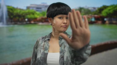 Arka planda yeşil ağaçlar olan açık hava parkında jest yapan bir kadın, su kenarında desenli gömlek giyen Çinli genç bir bayanı gösteriyor.