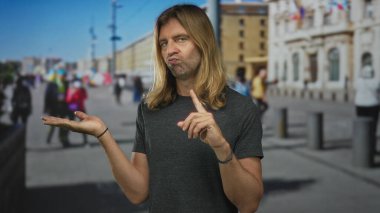 Açık avuç gösteren gri gömlekli bir adam güneşli bir caddede yukarı doğru işaret ediyor..