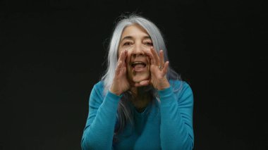 Gri saçlı kadın, siyah arka plana karşı anlamlı bir şekilde konuşuyor, mavi kazak giyiyor, izole bir iletişim ve ifade sahnesinde enerji ve duygu taşıyor..