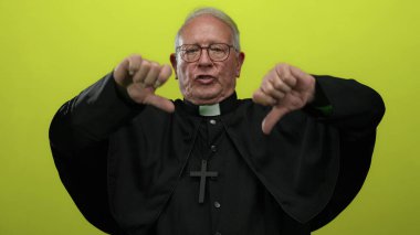 Siyah cüppeli kıdemli rahip, parlak sarı bir arka plana baş parmak işareti yaparak ciddi bir ifadeyle onaylamadığını iletiyor..