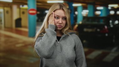 Sarı saçlı, gri kazaklı bir kadın kapalı bir otoparkta duruyor, yüzünde bir elle, arabalarla sarılmış bir şekilde..