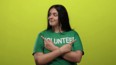Genç ve cüsseli gönüllü kadın, izole edilmiş sarı arka plan duvarına karşı yeşil gömlek giyerek ve kendinden emin bir ifade sergileyerek her iki tarafı da işaret ediyor..